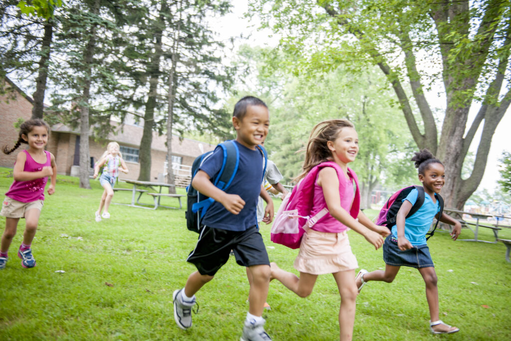 A diverse group of elementary-aged children wearing backpacks running joyfully across a lush green schoolyard on a sunny day.