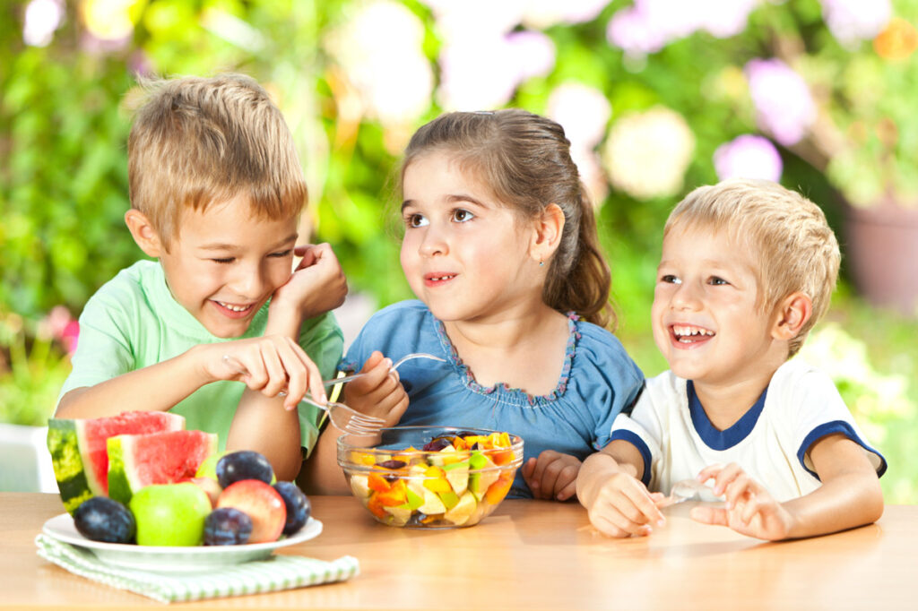 Three young children sitting at a wooden table outdoors, smiling and laughing while sharing fresh fruit. A plate of watermelon slices, plums, and a green apple sits in the foreground next to a large glass bowl of mixed fruit salad.