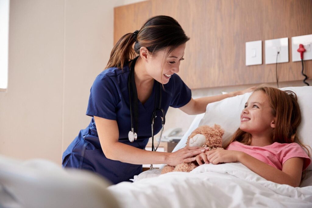 A smiling female nurse in blue scrubs leans over a young girl lying in a hospital bed giving best treatment