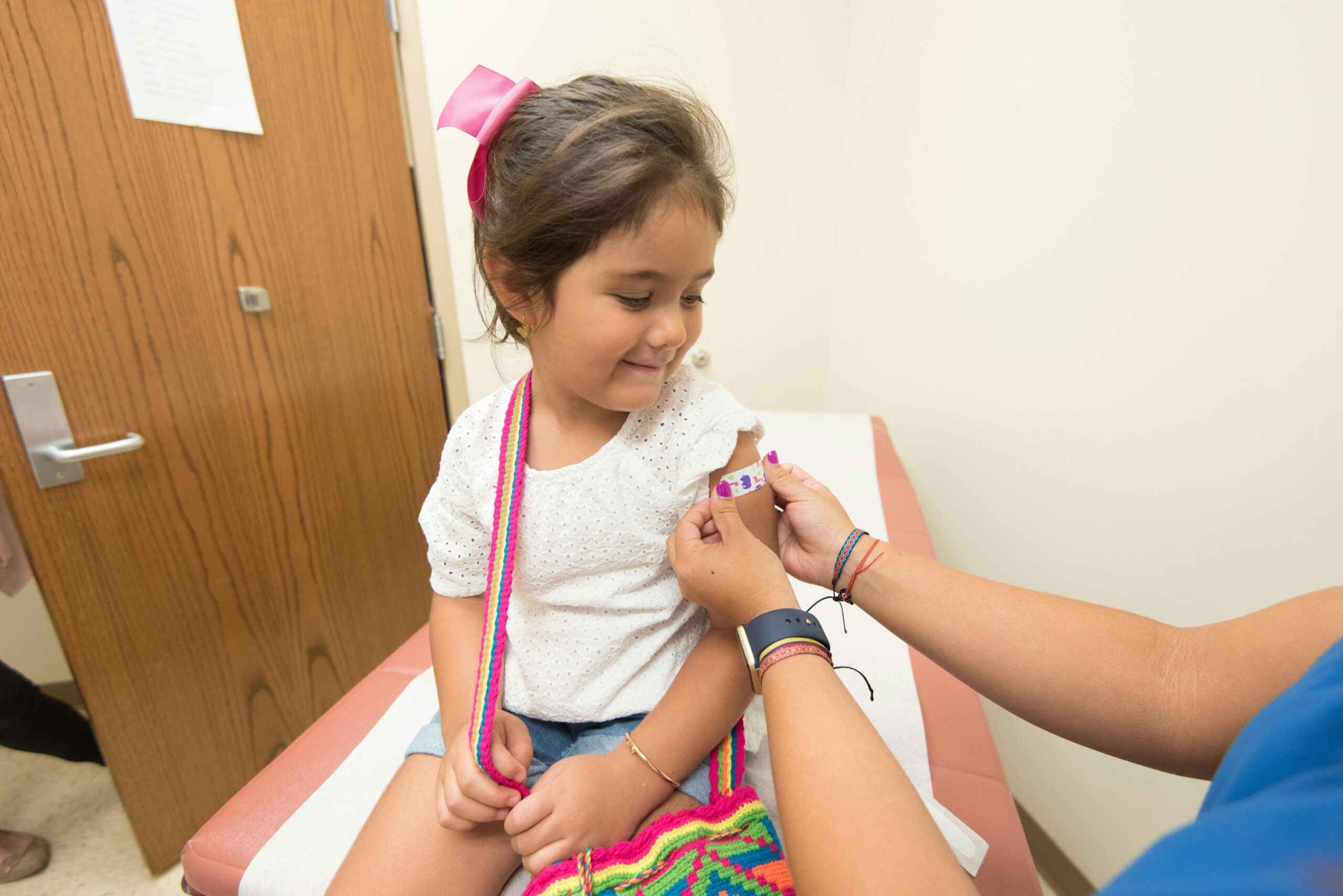 A young girl smiles as a healthcare worker places a bandage on her arm after a vaccination in a clinic room.