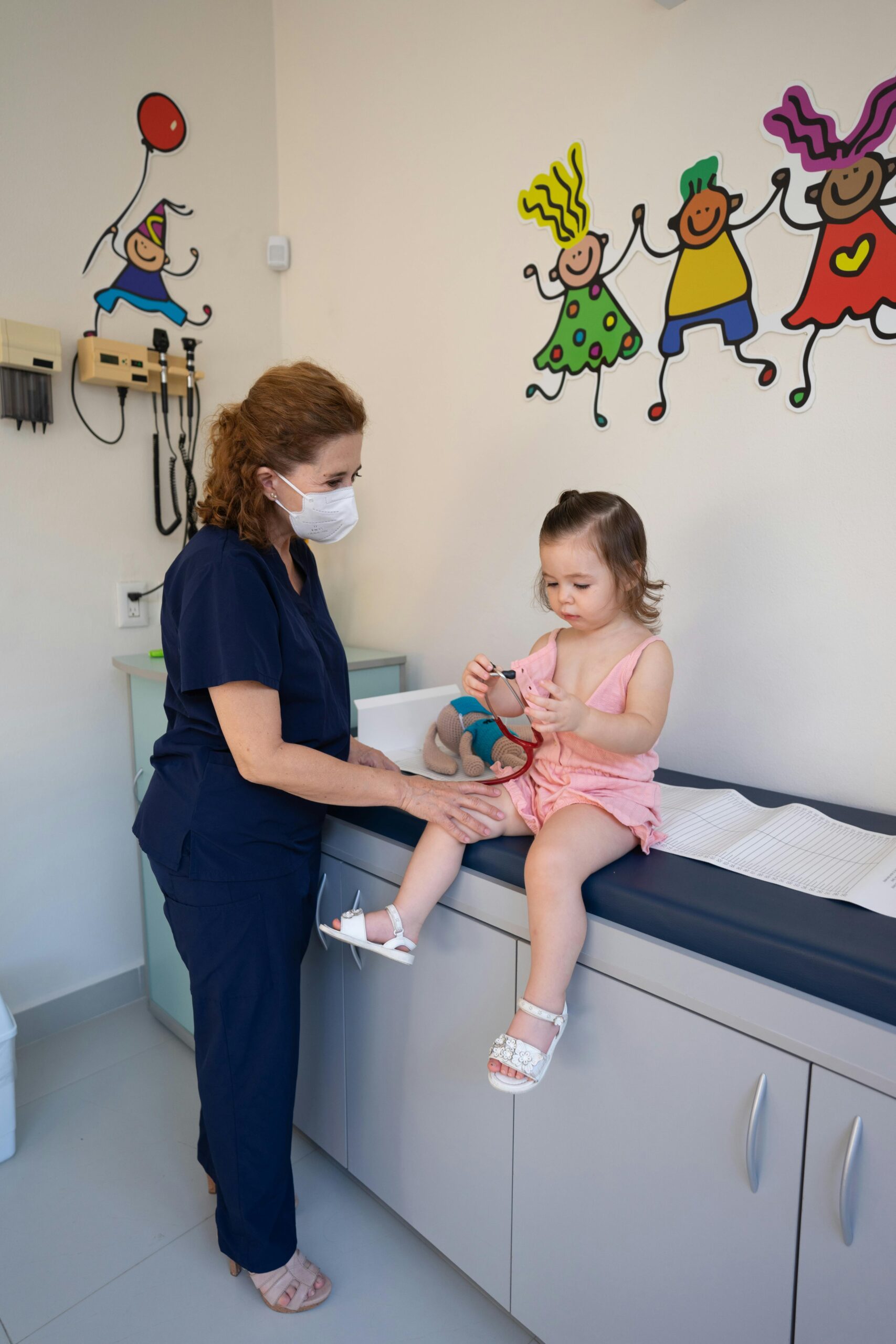 A nurse wearing a mask gently examines a young child sitting on an examination table in a colorful pediatric clinic room.